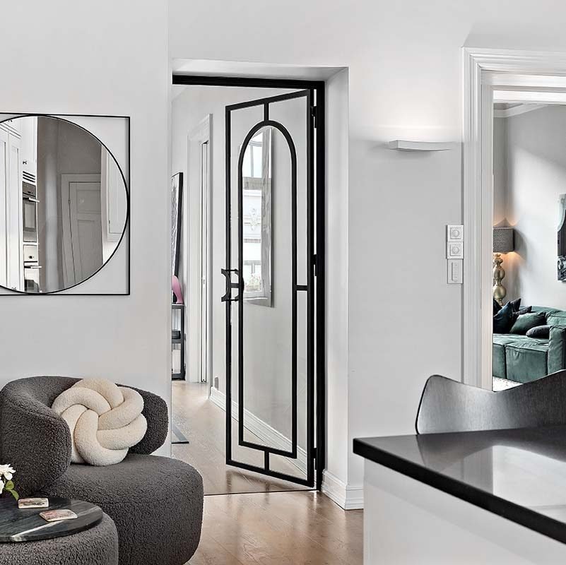 Hengslet glassdør med svart metallramme og buet toppdesign, plassert i et moderne interiør med lyse vegger, tregulv og minimalistisk innredning, inkludert en rund dekorativ speilramme og en grå loungestol